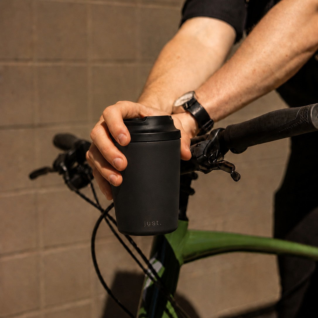 Person holding a black reusable coffee cup from Just Bottle on a bicycle.