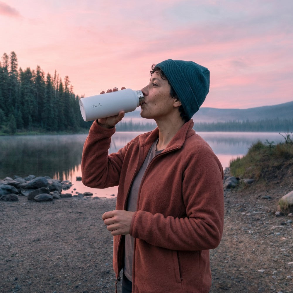 Person drinking from a Just Bottle by a lake at sunset