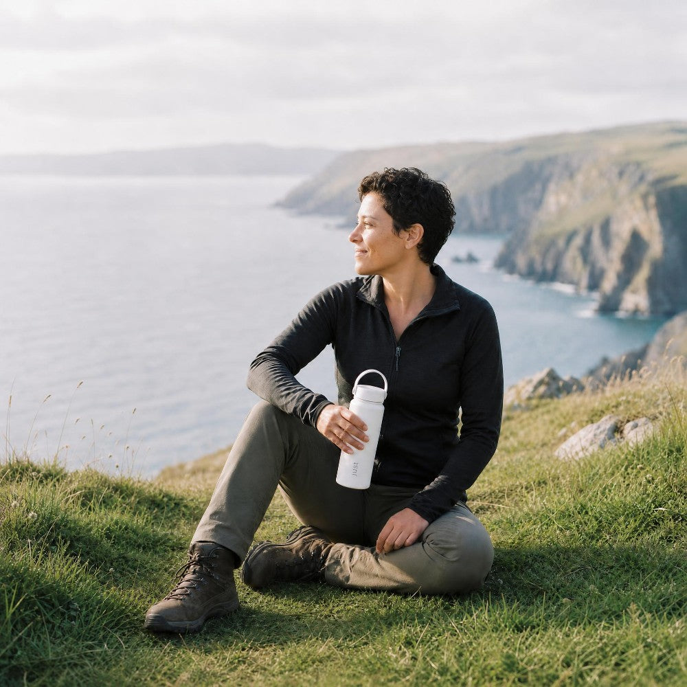 Person sitting on a grassy hill by a body of water holding a white 550ml stainless steel water bottle.