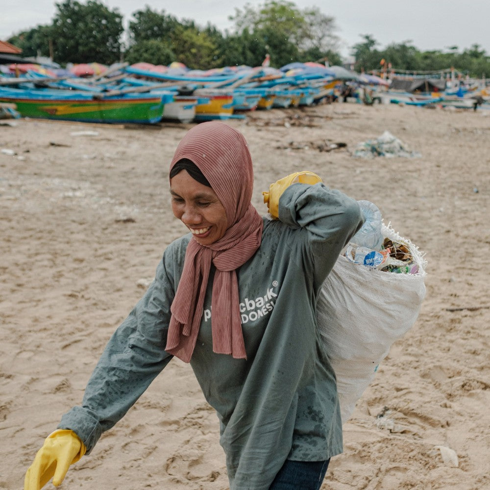 A smiling woman wearing a dusty pink hijab, a gray long-sleeve shirt with white text, and yellow gloves walks on a sandy beach while carrying a large white sack of plastic bottles on her back. Colorful boats and trees are visible in the background.