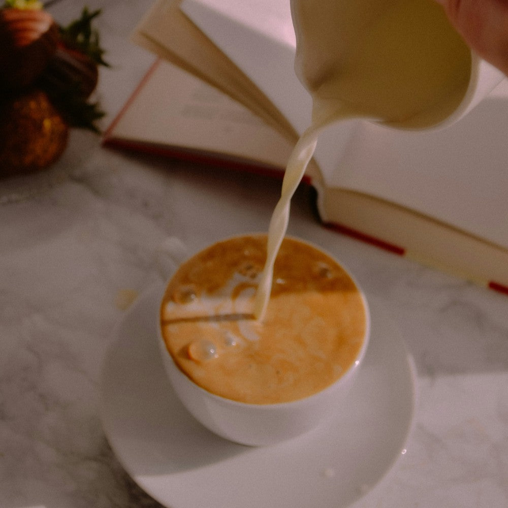Pouring milk into a cup of coffee on a marble surface with a book in the background.