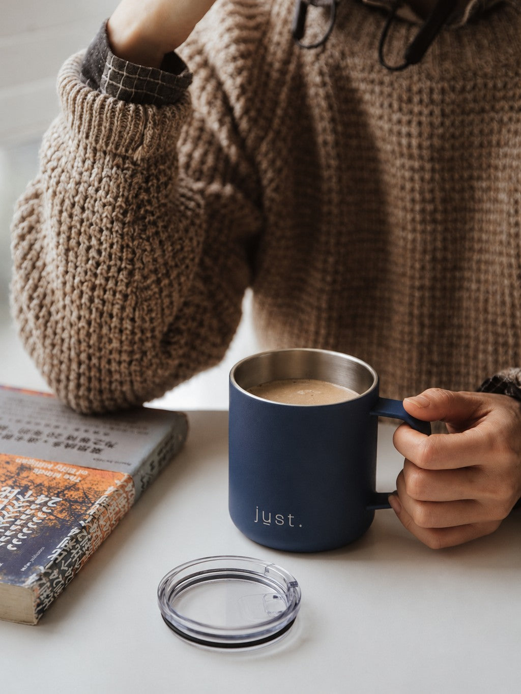 Person holding a Just Bottle 10oz dark blue camp mug, wearing knitted sleeves, on a light surface with a book and lid.