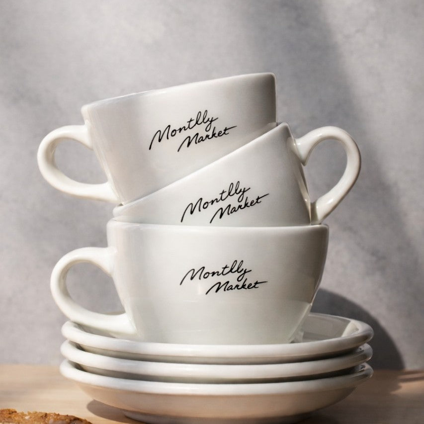 Stack of white ceramic 150ml cafe cups and saucers with 'Monthly Market' text on a neutral background