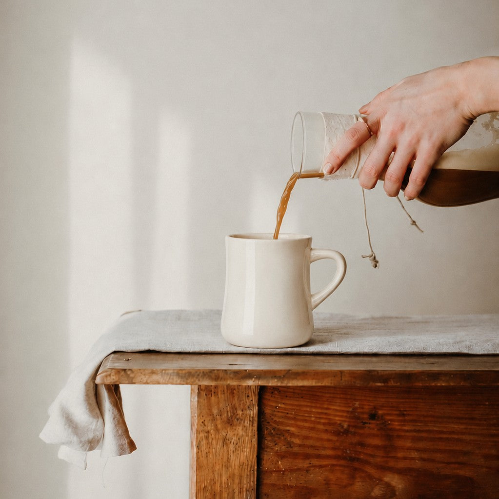 Person pouring cold brew coffee from a glass container into a white classic diner mug on a wooden surface.