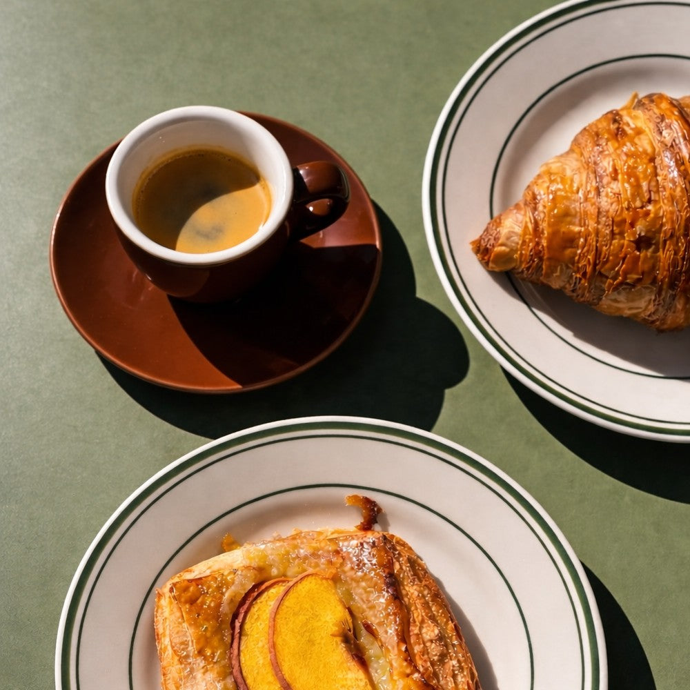 Just Bottle porcelain espresso cup and saucer with a croissant and a pastry on plates against a green background