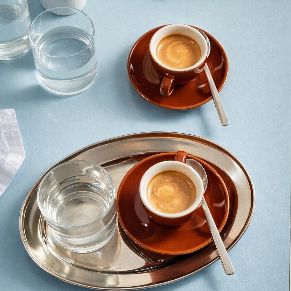 Two Just Bottle Origin 75ml espresso cups on brown saucers with spoons, placed on a metal tray against a light blue background.
