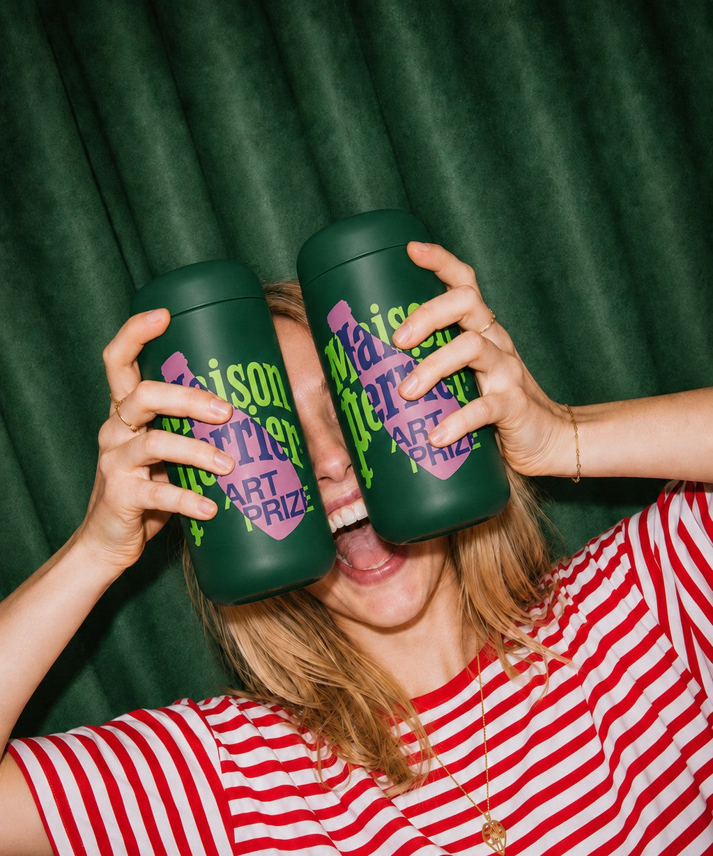 A woman in a red and white striped shirt holds two dark green leakproof tumblers with custom 'Maison Perrier' graphics in front of her eyes, set against a dark green curtain background.