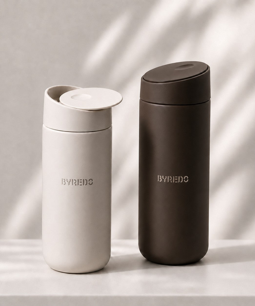 Two custom branded stainless steel travel tumblers with slide top lids, one in light warm gray and one in dark brown, standing side by side on a light surface with soft shadow patterns.