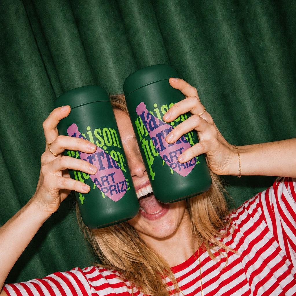 A woman in a red and white striped shirt holds two dark green leakproof tumblers with purple and lime green text reading 'Maison Perrier ART PRIZE' in front of her eyes, against a dark green curtain background.