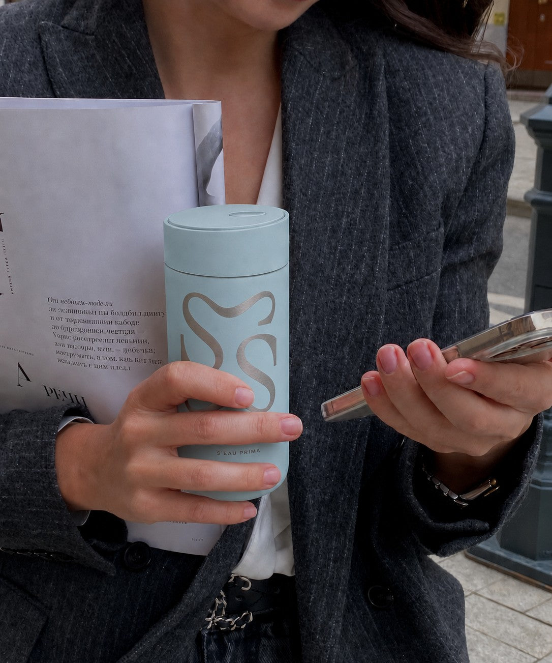 A close-up lifestyle shot of a person holding a light blue travel tumbler with a laser etched logo. The person is wearing a dark gray blazer and holding a white book in one hand and a smartphone in the other.