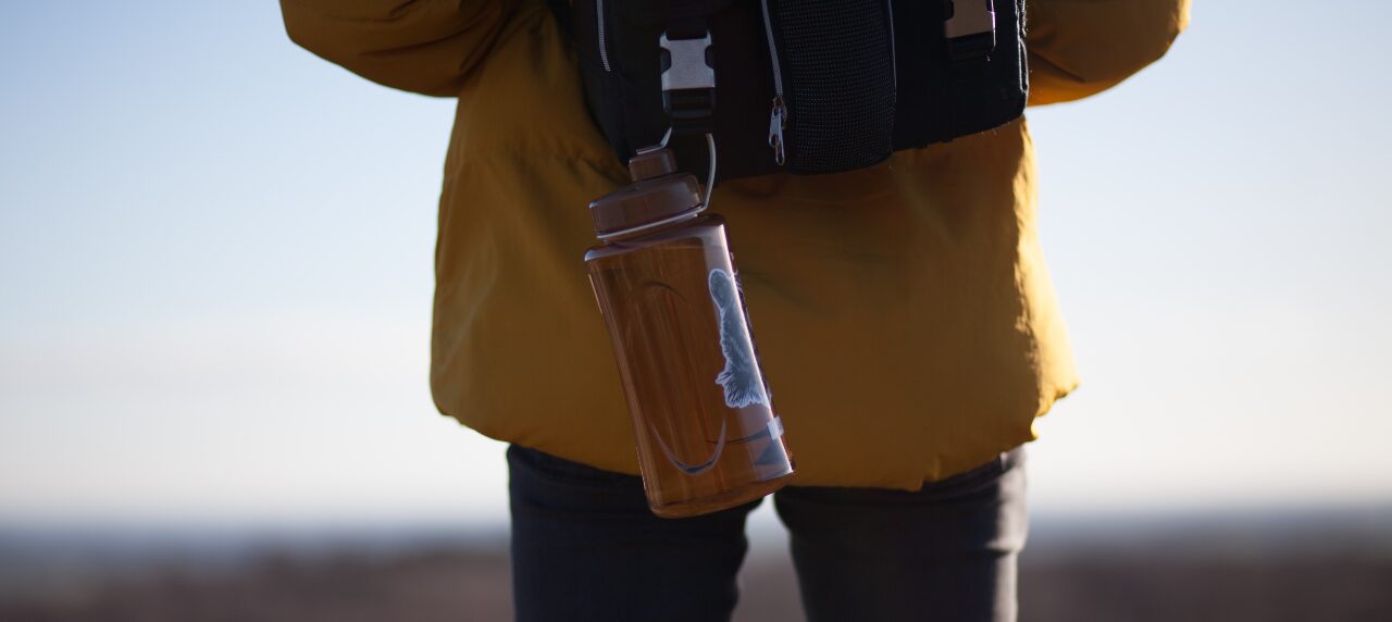 Glass water bottle hanging from backpack with nature behind