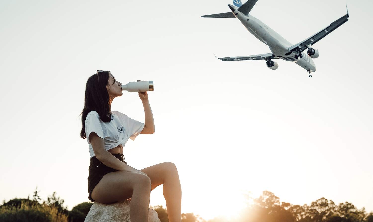 lady drinking water from reusable bottle with sunset and aircraft flying in background