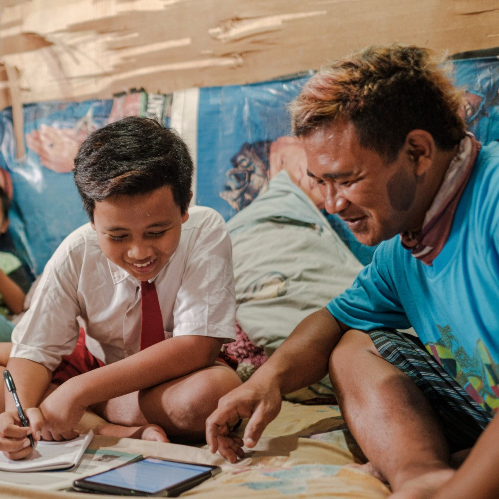A smiling young boy in a white shirt and red tie sits beside an adult man in a bright blue t-shirt, looking at a tablet together in a simple indoor setting.