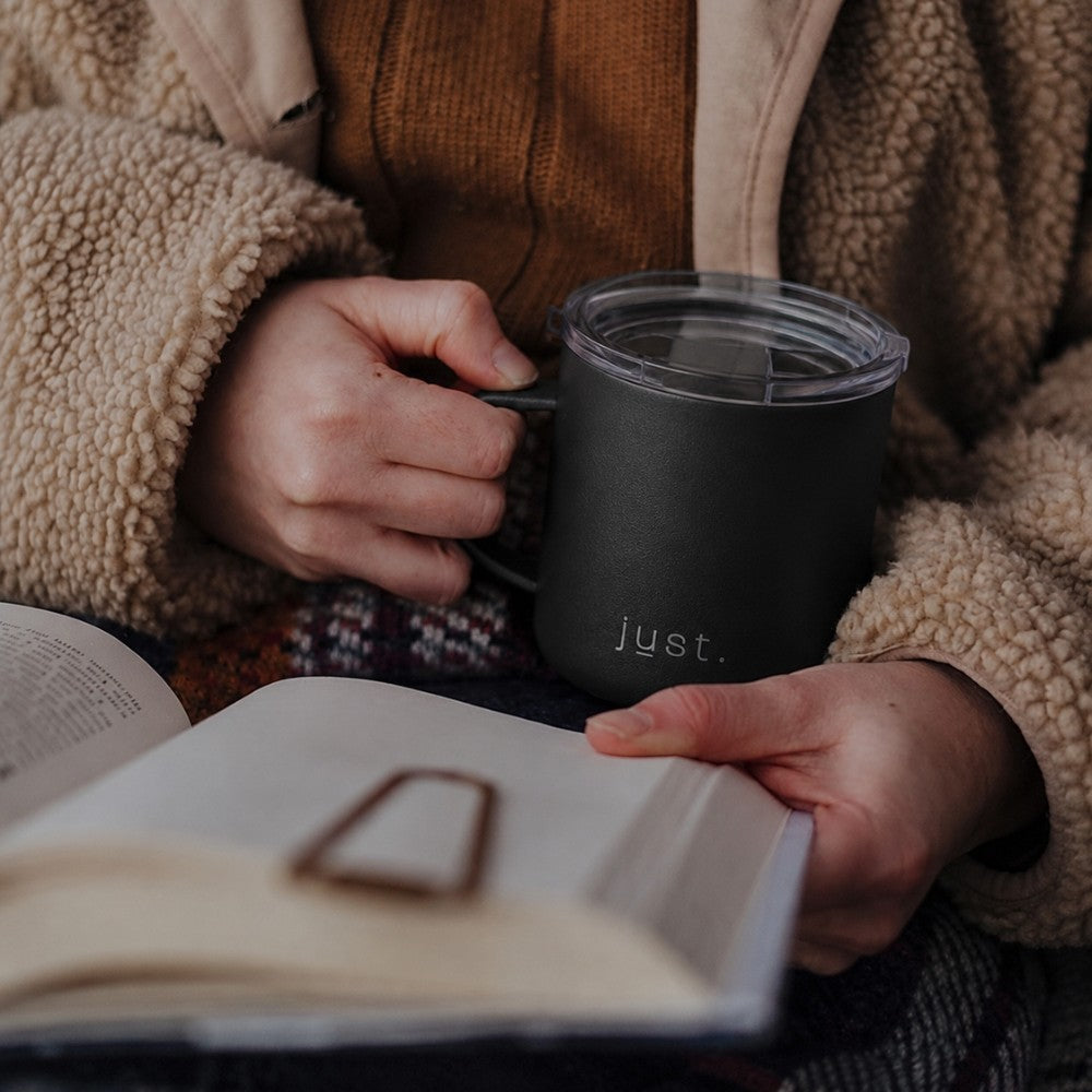 Person holding a black metal camp mug labeled from Just Bottle and an open book, wearing a brown jacket.