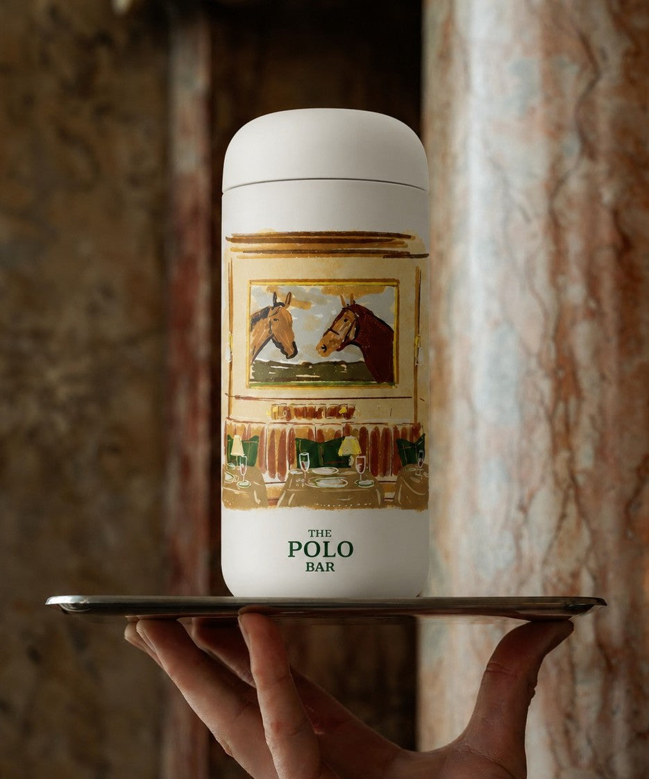 A white leakproof tumbler, featuring a custom print for "The Polo Bar." The tumbler is held on a metallic tray by a hand against a warm marble background.