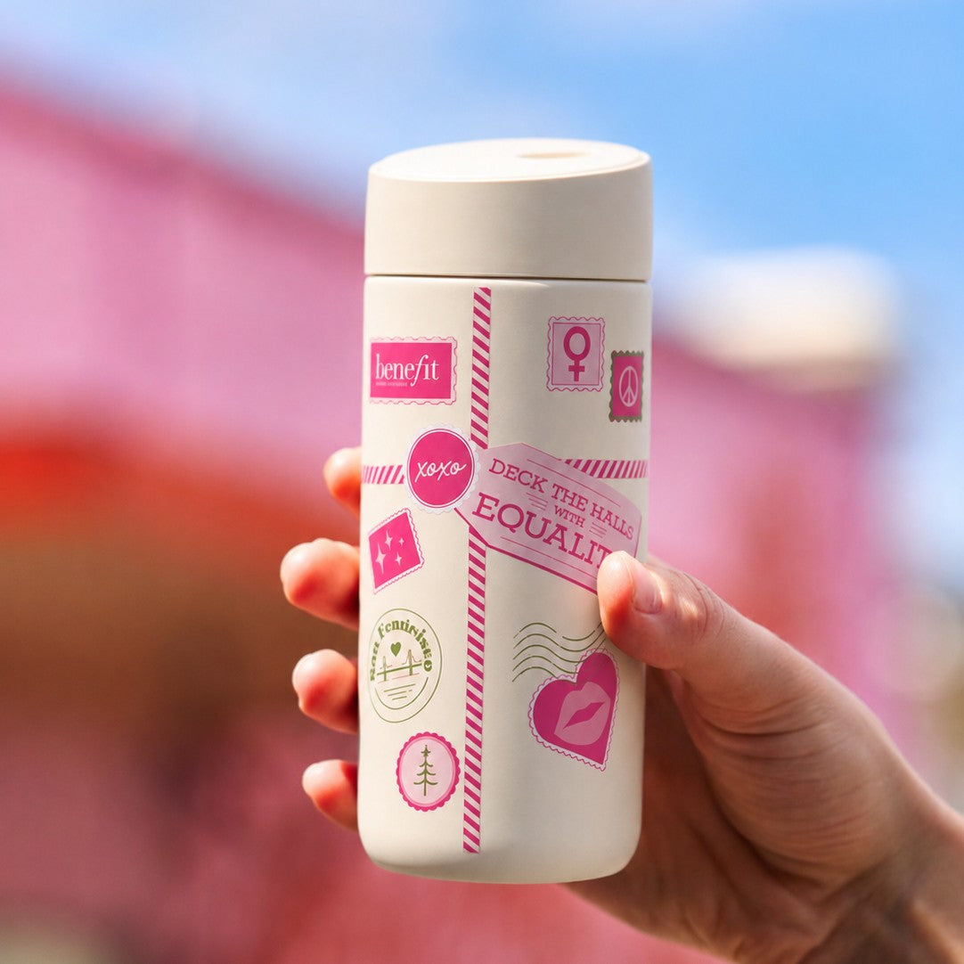 A hand holding a white metal travel tumbler with pink and multicolor graphic logos, set against a blurred pink and blue background.