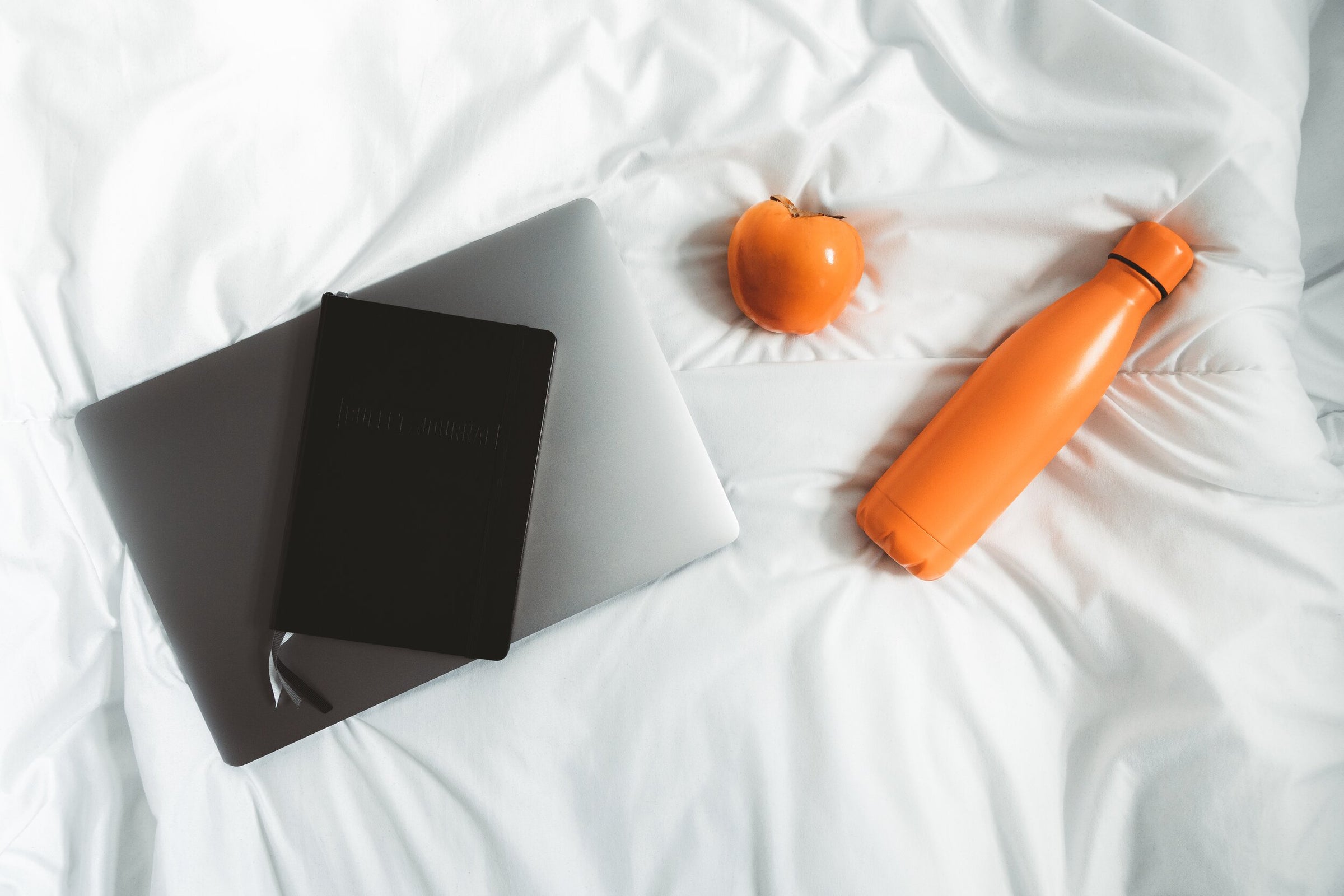orange reusable water bottle next to fruit and a laptop on a bed