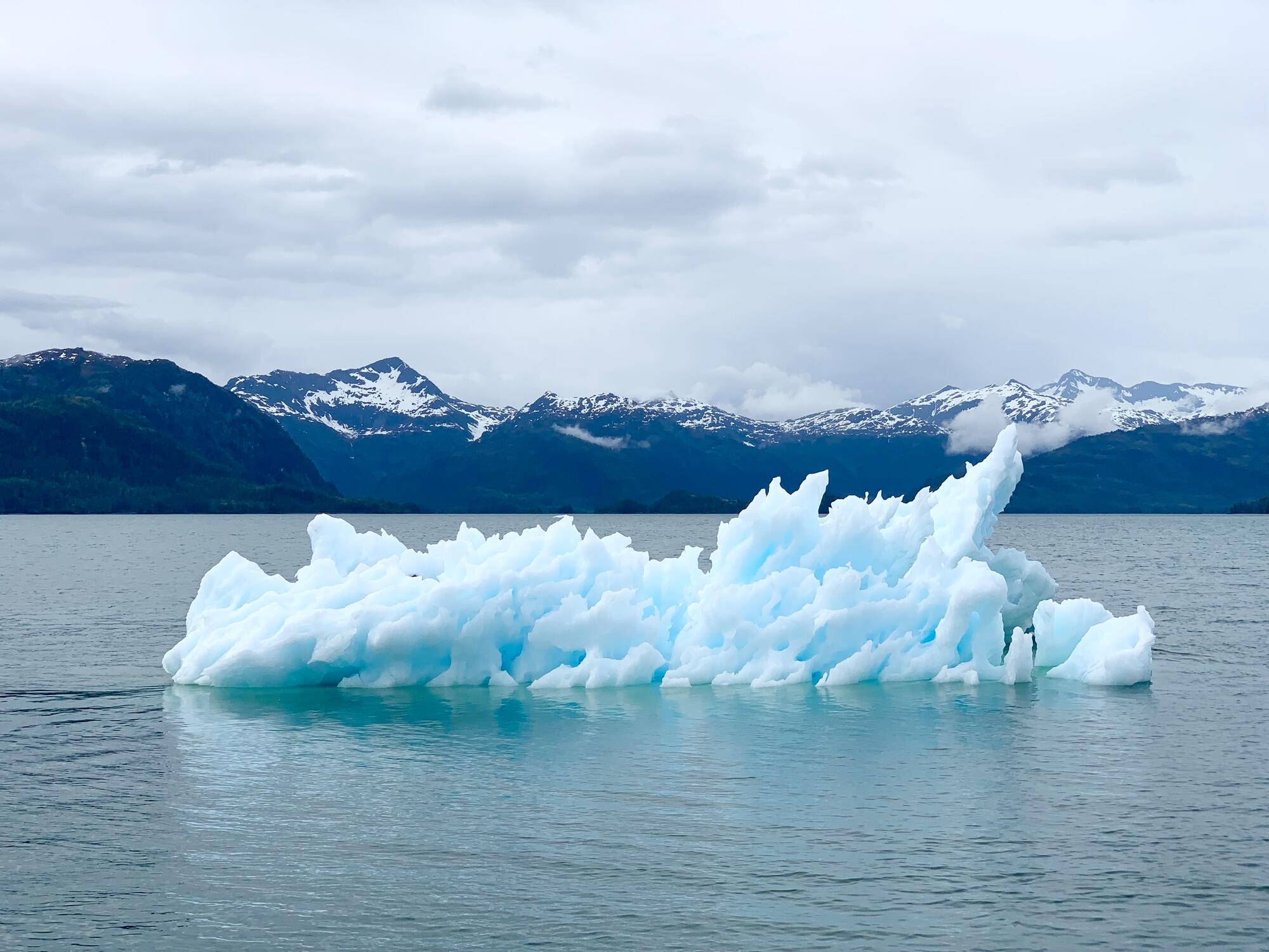 small iceberg floating in ocean in front of snow capped mountains 