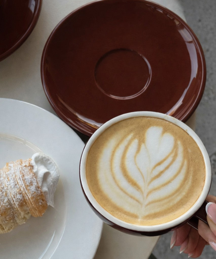 Cup of coffee with latte art, pastry, and saucer on a white surface