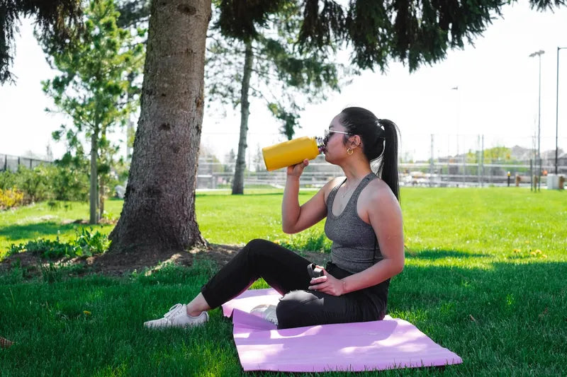 lady drinking from a yellow water bottle sitting on a yoga mat under a tree